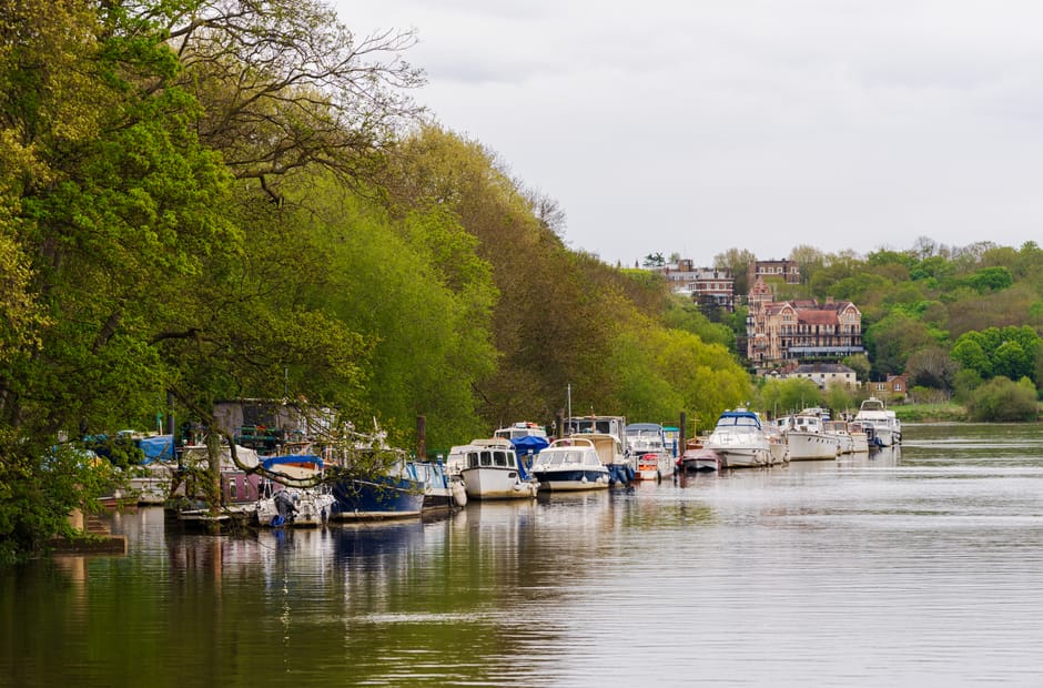 A wide-angle view of a river in Richmond upon Thames during early evening, with the sky showing dark, cloudy weather. Several boats of various sizes and colors are moored along the riverbank, some cov