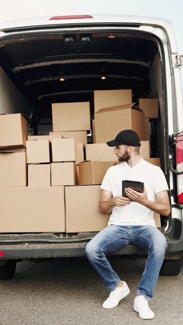 A man with a beard, wearing a black cap, white t-shirt, and blue jeans, sits on the edge of an open vehicle rear door. Inside the vehicle, numerous cardboard boxes of various sizes are stacked, some w