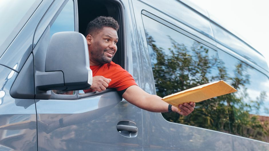A man with a beard, wearing a black cap, white t-shirt, and blue jeans, sits on the edge of an open vehicle rear door. Inside the vehicle, numerous cardboard boxes of various sizes are stacked, some w