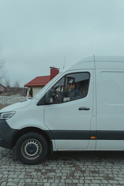 A man wearing a dark vest, black pants, and a cap is standing outside next to an open white van with rear doors swung open on a paved surface. The van is fully loaded with multiple cardboard boxes of