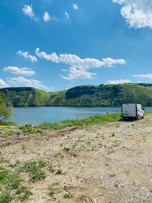 A white van parked on a gravel driveway at the edge of a river or lake with green hills and cliffs in the background under a partly cloudy blue sky. In the foreground, the gravel surface has sparse grass and small plants. The van is positioned near a building or property entrance, with the side door closed, and is ready for loading or unloading. This setting illustrates a scenic location suitable for home relocation or furniture transport services, potentially requiring careful packing and logistics management as part of a moving operation with Man with Van Mortlake, offering professional removals and moving services nearby.