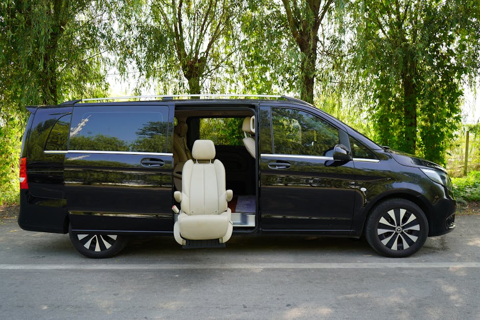 A black van with its side sliding door open, parked on a paved road with green trees and foliage in the background. The van’s interior is visible, showing beige leather seats and various packed moving boxes made of cardboard with plastic wrapping, arranged inside for a home relocation. One beige captain’s chair is partially extended outside the van, ready for loading or unloading, indicating a furniture transport process. The vehicle is situated on a flat surface in natural daylight, suggesting an outdoor moving operation near the Mortlake Riverside flats on Lower Richmond Road, where [COMPANY_NAME] provides removals services. Trolley straps and blankets may be seen securing or protecting the furniture during the loading process, with the surrounding environment emphasizing the accessibility for van access as described in the page titled 'Mortlake Riverside flats: van access on Lower Richmond Rd, MORTLAKE'.