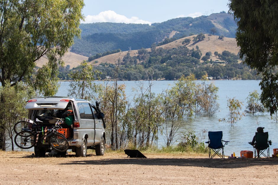 A white van parked on a gravel driveway at the edge of a river or lake with green hills and cliffs in the background under a partly cloudy blue sky. In the foreground, the gravel surface has sparse grass and small plants. The van is positioned near a building or property entrance, with the side door closed, and is ready for loading or unloading. This setting illustrates a scenic location suitable for home relocation or furniture transport services, potentially requiring careful packing and logistics management as part of a moving operation with Man with Van Mortlake, offering professional removals and moving services nearby.
