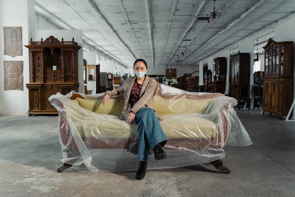 A woman wearing a face mask, a beige checked blazer, and blue jeans is seated on a large, antique-style sofa inside a spacious and industrial-looking warehouse. The sofa is partially wrapped in protective bubble wrap for transport, with visible plastic covering the fabric upholstery and wooden legs. Surrounding her are various pieces of vintage wooden furniture, including cabinets and display units, arranged along the warehouse walls. The ceiling features exposed concrete and industrial lighting fixtures, and the floor is concrete, indicating a site used for home relocation and furniture transport. The woman appears to be in the process of a furniture move or inspection, representing the careful handling involved in specialist removal services by Man with Van Mortlake, who manage packing, moving, and loading of antique and valuable items during residential relocations.