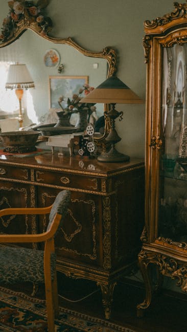 A woman wearing a face mask, a beige checked blazer, and blue jeans is seated on a large, antique-style sofa inside a spacious and industrial-looking warehouse. The sofa is partially wrapped in protective bubble wrap for transport, with visible plastic covering the fabric upholstery and wooden legs. Surrounding her are various pieces of vintage wooden furniture, including cabinets and display units, arranged along the warehouse walls. The ceiling features exposed concrete and industrial lighting fixtures, and the floor is concrete, indicating a site used for home relocation and furniture transport. The woman appears to be in the process of a furniture move or inspection, representing the careful handling involved in specialist removal services by Man with Van Mortlake, who manage packing, moving, and loading of antique and valuable items during residential relocations.