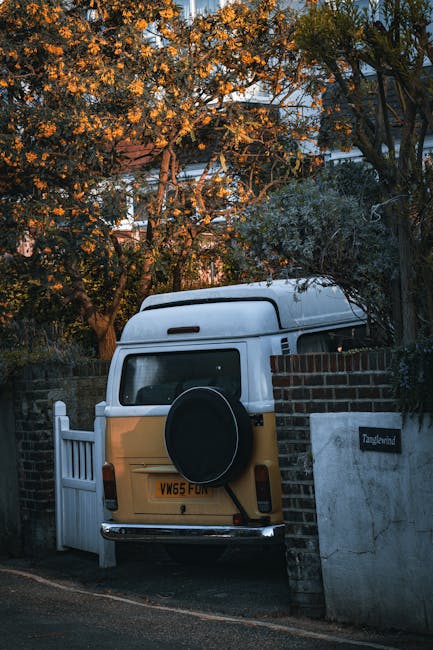 A vintage Volkswagen camper van with a yellow lower half and white upper section is parked on a narrow residential street next to a brick wall and a white picket gate. The van is positioned closely to the residential property, with its rear door facing outward, and a spare tire mounted on the back. The street is lined with trees that have autumn-colored leaves, partially illuminated by warm evening sunlight. The surrounding environment includes leafy trees, a brick wall, and a small nameplate reading 'Tanglewood' on the brick wall next to the van. This scene reflects a typical residential area suitable for house removals and home relocation services by Man with Van Mortlake, highlighting the careful loading and parking process involved in furniture transport and packing during a move.