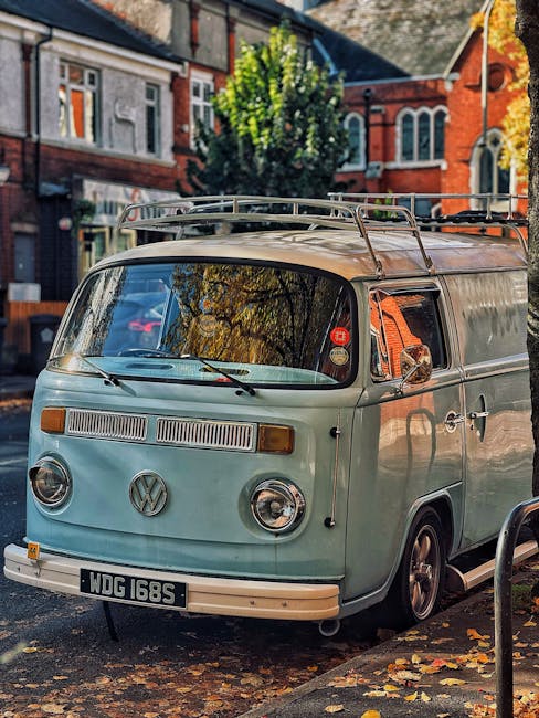 A blue vintage panel van is parked on a paved pavement alongside a black metal fence in front of a large brick wall. The van is positioned close to the curb, casting a shadow on the ground, and appears to be ready for a home relocation or furniture transport as part of a moving service. The brick wall behind the van is uniform in color with some visible horizontal and vertical mortar joints, and there are no windows or visible building entrances on this section of the wall. Overhead, a single wire runs horizontally across the image, attached to the wall with small fixtures. The lighting indicates daytime with natural sunlight illuminating the scene evenly, highlighting the texture of the brickwork and the smooth surface of the van. This setting reflects a typical loading or unloading area associated with house moving, and the image aligns with the professional relocation services offered by Man with Van Mortlake in SW14, focusing on efficient logistics and furniture transport during home moves.