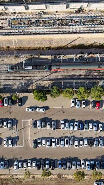 An aerial view showing a railway track and platform above a parking lot filled with numerous parked cars in various colours, including silver, white, red, and blue. A train with a dark grey body and blue and red detailing is positioned on the tracks, with no visible passengers or crew. The parking area features marked spaces with white lines, and a few small trees are scattered along the edges. The environment appears urban, with the sunlight casting shadows of the cars and trees onto the asphalt. This scene illustrates the logistical context of house removals and furniture transport, highlighting the importance of planning for loading and parking when conducting home relocation near Mortlake Station, as managed by Man with Van Mortlake.