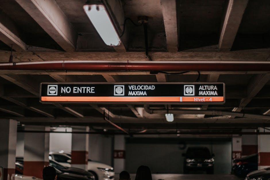 Inside an underground parking garage with a low ceiling featuring exposed concrete beams and a fluorescent ceiling light. The ceiling contains pipes and wiring visible above a digital sign displaying parking restrictions in Spanish, indicating a maximum speed limit of 10 km/h and a maximum height clearance of 4 metres. The sign also states 'NO ENTRE' in bold, black letters on a white background. Below the sign, vehicles are parked along the concrete floor, and some individuals are visible in the lower part of the image, suggesting active loading or movement. The environment is typical for urban parking areas next to residential or commercial buildings, where house removals or furniture transport could be organized, making it relevant to the services of Man with Van Mortlake in facilitating home relocation and loading processes near Mortlake Station.