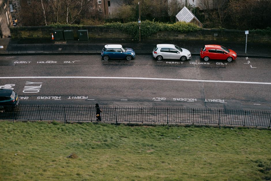 An aerial view of a street scene showing three parked cars on a permit parking zone marked with white painted lines and text that reads 'PERMIT HOLDERS ONLY' and 'TWO HOUR LIMIT'. The first car, a blue compact vehicle with a white rooftop cargo box, is positioned closest to the sidewalk. The second car, a silver hatchback, is parked beside it, and the third, a red hatchback, is located further along the curb. A pedestrian dressed in dark clothing stands near the black metal fence separating the parking area from a grassy field in the foreground. The grassy area appears well-maintained with a few small patches of uneven growth. In the background, there are trees, shrubs, and a building partially obscured by foliage, along with some green waste bins aligned along the sidewalk. The scene is illuminated by natural daylight, capturing a typical urban environment suited for packing, home relocation, and furniture transport activities, which are often coordinated by professional removals companies like Man with Van Mortlake during scheduled moving days.