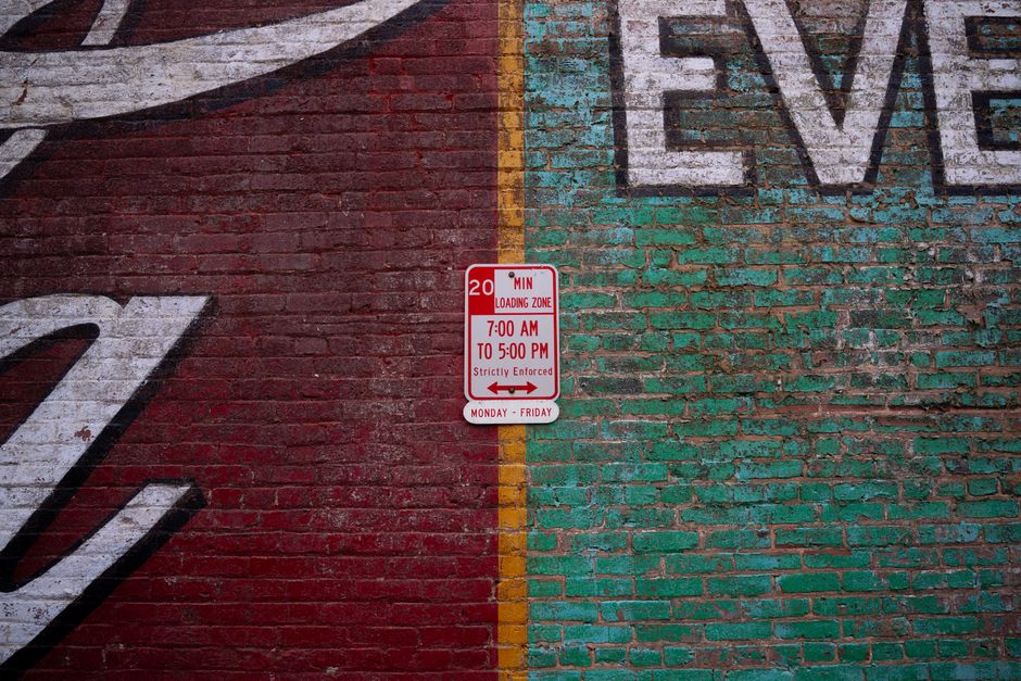 A red and white parking regulation sign affixed to an aged brick wall, indicating a loading zone from 7:00 AM to 5:00 PM, Monday to Friday, with a 20-minute limit. The wall features a painted mural or graffiti, with the word 'LOVE' partially visible in large, capital letters painted in faded white and blue. A yellow vertical line runs through the middle of the brick surface, dividing the painted sections. The area appears to be outdoors, likely on a street in Mortlake, with natural lighting highlighting the textures of the bricks and the vibrant colours of the sign and mural. This setting reflects urban environments where residents and professional movers, such as those from Man with Van Mortlake, may arrange for loading and unloading during home relocation or furniture transport activities.