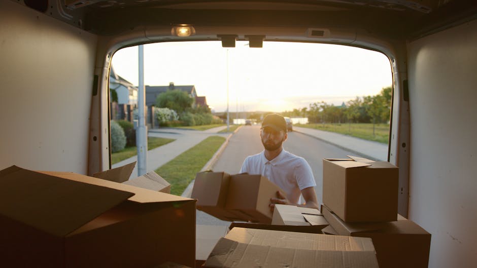 A man wearing a white polo shirt and glasses is standing outside a large moving van during a home relocation, with the van's open rear door revealing the interior filled with several cardboard boxes and packing materials. The boxes vary in size, some are plain brown cardboard, while others are sealed with packing tape, and a few are stacked on top of each other near the van's opening. The man is holding a medium-sized box, preparing for loading onto the van, which is situated on an urban street with curb sidewalks, residential houses, and greenery in the background. The scene is illuminated by warm, natural sunlight, suggesting late afternoon or early evening, and the overall setting reflects the process of furniture transport and packing during a professional removal or moving service, as provided by Man with Van Mortlake, supporting secure and organized house removals, including the necessary permits for moving vans in Richmond Council area.