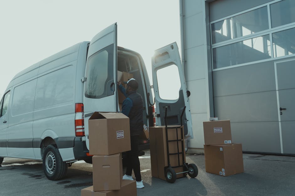 A mover from Man with Van Mortlake is loading cardboard boxes and packing materials into a white commercial van, parked outside a modern building with large glass windows. The boxes are of various sizes, some sealed with packing tape and labelled for moving, and are arranged on the pavement alongside a black hand truck or trolley, which is being used to transport larger items. The mover is wearing casual work attire, including a dark jacket and a cap, and is seen lifting or placing boxes into the vehicle's cargo area, which has its rear door open. The scene captures the initial stages of a home relocation or furniture transport, with a focus on packing, loading, and organizing items for transport. The lighting suggests daylight with sunlight slightly behind the van, creating a bright outdoor environment suitable for detailed visual documentation of the loading process. Man with Van Mortlake operates within this context, supporting secure and efficient moving services, including obtaining necessary permits for van activities in residential areas such as Mortlake.