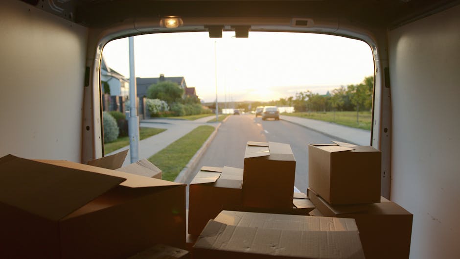 A man wearing a white polo shirt and glasses is standing outside a large moving van during a home relocation, with the van's open rear door revealing the interior filled with several cardboard boxes and packing materials. The boxes vary in size, some are plain brown cardboard, while others are sealed with packing tape, and a few are stacked on top of each other near the van's opening. The man is holding a medium-sized box, preparing for loading onto the van, which is situated on an urban street with curb sidewalks, residential houses, and greenery in the background. The scene is illuminated by warm, natural sunlight, suggesting late afternoon or early evening, and the overall setting reflects the process of furniture transport and packing during a professional removal or moving service, as provided by Man with Van Mortlake, supporting secure and organized house removals, including the necessary permits for moving vans in Richmond Council area.