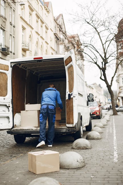A professional removal technician wearing a blue jacket and dark trousers is seen loading a large cardboard box into the open rear of a white moving van parked on a city street. Inside the van, several stacked cardboard boxes and padded furniture blankets are visible, indicating packing and furniture transport for a home relocation. The van is positioned next to a curb with concrete bollards separating the street from the pavement. Surrounding the scene are multi-storey residential buildings with classic architectural features, including large windows and ornate facades, and leafless trees indicating a colder season. Additional parked cars line the street, and the overall environment suggests a busy urban area prepared for a same-day removals service, as offered by Man with Van Mortlake, who specializes in rapid and efficient moving and logistics solutions.