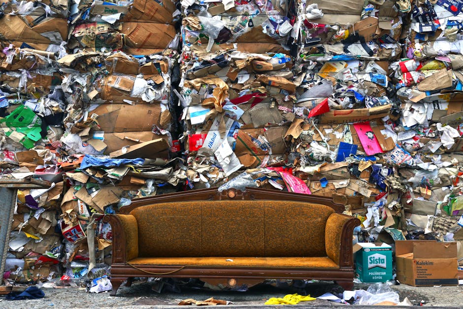 A vintage wooden and upholstered sofa with a curved wooden frame and orange-brown fabric is positioned on the pavement in front of a large, densely packed pile of compressed cardboard and paper waste. The waste includes flattened cardboard boxes, crumpled paper, and packaging materials in various colors such as brown, white, red, blue, and green, some with printed logos and text. The pile extends across the background, partially obscuring a building or structure behind it. The scene is outdoors, with natural daylight illuminating the waste and furniture, illustrating a situation where proper disposal or removal services like those offered by Man with Van Mortlake are needed for home relocation or furniture transport, especially when disposing of bulky items without council fines.