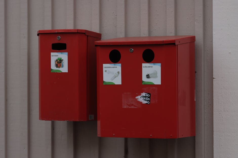 Two red postboxes mounted on a vertical wooden wall, with a slightly textured surface and vertical paneling. The postbox on the left is a narrow, rectangular shape with a slot for mail at the top and a small label featuring a battery illustration underneath. The larger, adjacent postbox on the right has a sloped top and displays two circular slots near the top, each covered with black circular openings. Below these slots, there are two paper labels showing images of lightbulbs, one with a filament lightbulb labeled 'LAMPENGLAMPOR' and the other with an LED bulb labeled 'GLÖDLAMPOR', attached with a transparent adhesive. The image appears captured outdoors under natural lighting, possibly near a residential or commercial property, with the focus on the postboxes commonly used in house removals for collecting mail or disposing of bulky items. The setting emphasizes the process of packing or preparing for a move, with the nearby external environment representing typical relocation logistics. Man with Van Mortlake occasionally ties into the scene's context, supported by the mention of house and furniture transport services.