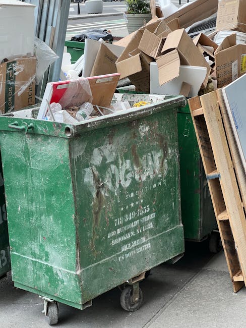 A vintage wooden and upholstered sofa with a curved wooden frame and orange-brown fabric is positioned on the pavement in front of a large, densely packed pile of compressed cardboard and paper waste. The waste includes flattened cardboard boxes, crumpled paper, and packaging materials in various colors such as brown, white, red, blue, and green, some with printed logos and text. The pile extends across the background, partially obscuring a building or structure behind it. The scene is outdoors, with natural daylight illuminating the waste and furniture, illustrating a situation where proper disposal or removal services like those offered by Man with Van Mortlake are needed for home relocation or furniture transport, especially when disposing of bulky items without council fines.